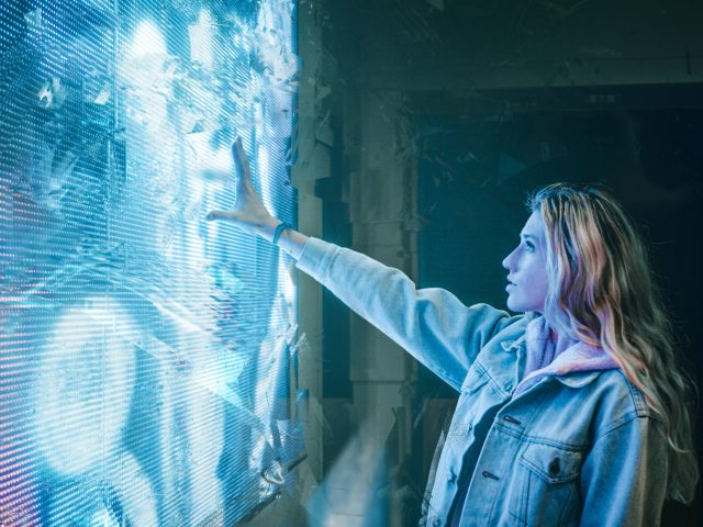 Eine Frau steht vor einem großen digitalen Bildschirm und streckt die Hand danach aus.